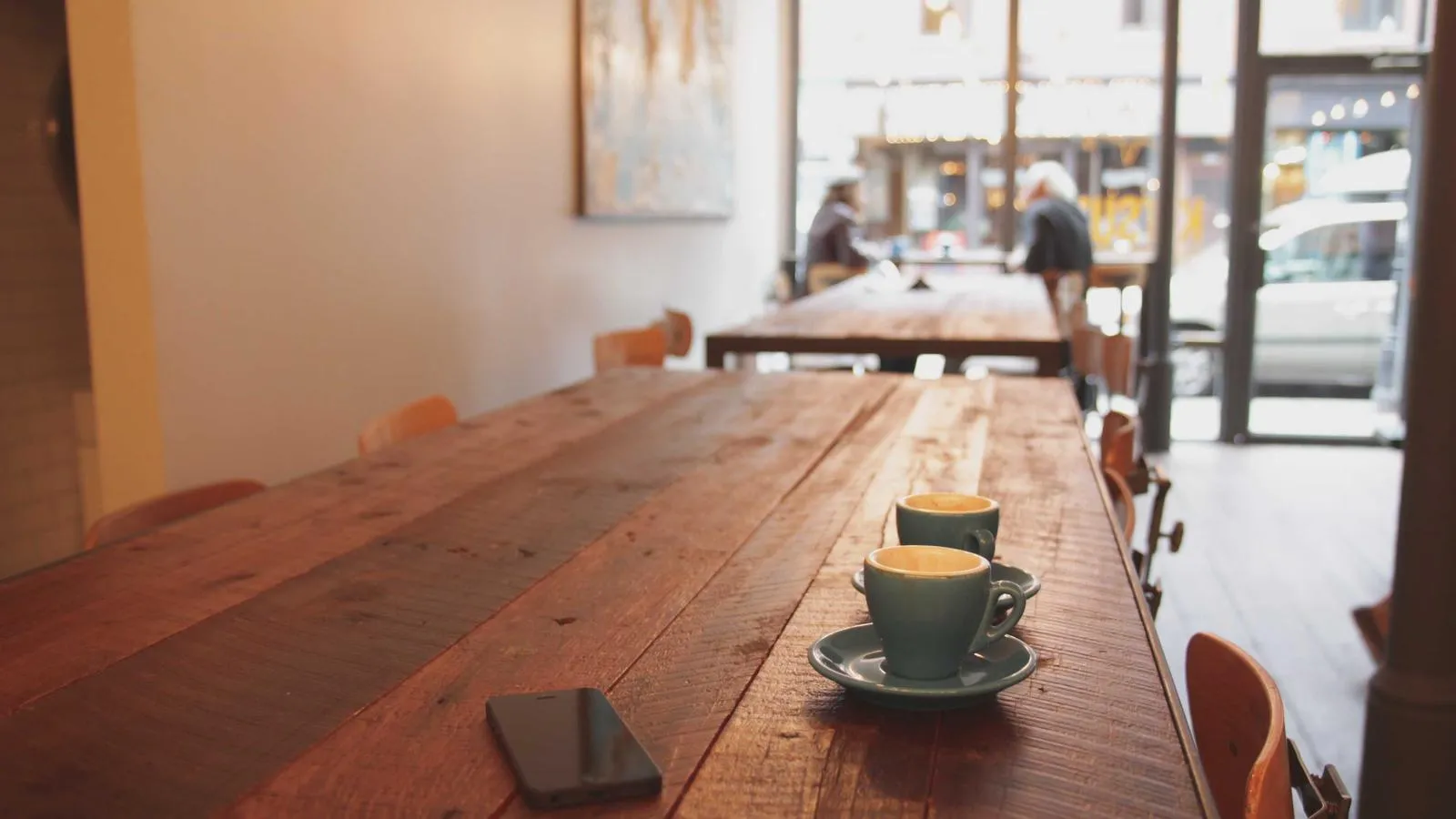 Two coffee cups on a long wooden table inside a sunlit café